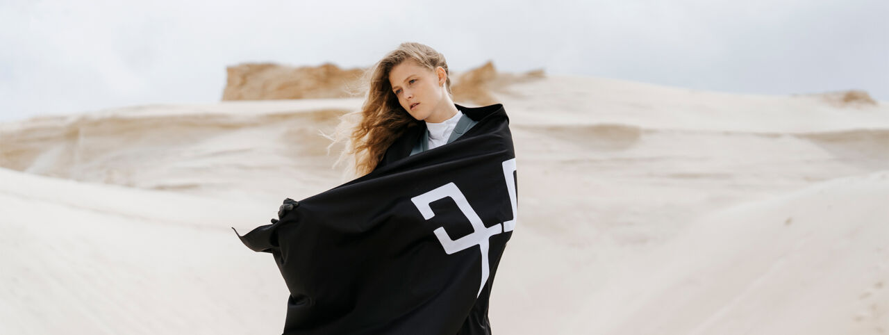 Dans un desert de sable blanc, une cavalière déterminée, tient un drapeau pour étendard. Sur son immense drapeau noir, repose l'emblème "horse pilot".